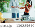 Renewable energy concept: wind turbine model, modern house miniature, and hard hat on desk with architects discussing sustainable design in blurred background. 139285640