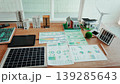 Desk workspace with solar panel, wind turbine model, smart home miniatures and printed energy analytics charts, illustrating renewable energy planning and sustainable city development. 139285643