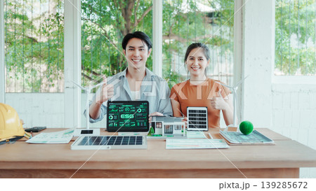 Two young professionals at desk giving thumbs up with laptop showing esg dashboard, solar panels, model house and documents, promoting sustainable energy and green building in bright office. Two young professionals at desk giving thumbs up with laptop showing esg dashboard, solar panels, model house and documents, promoting sustainable energy and green building in bright office. 139285672