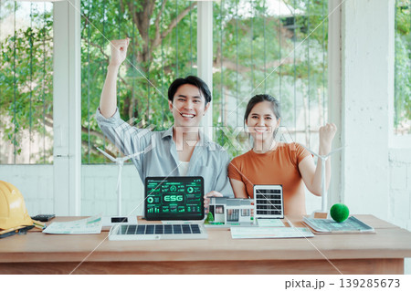 Smiling young team celebrates sustainable energy project at desk with laptop showing ESG dashboard, wind turbines and solar panel models in bright office with green view. Smiling young team celebrates sustainable energy project at desk with laptop showing ESG dashboard, wind turbines and solar panel models in bright office with green view. 139285673