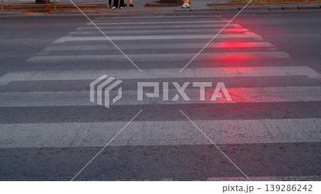 A crosswalk on a street with some people nearby. Street lights are on, casting a red glow 139286242