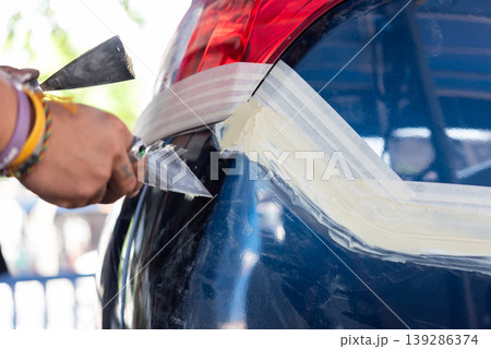 Close up of technician applying body filler on car panel with putty knife masking tape outlining repair area auto maintenance scratch dent restoration process in workshop 139286374