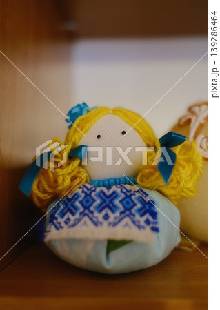 Doll wearing a blue and white patterned dress sits upright on a brown wooden shelf 139286464