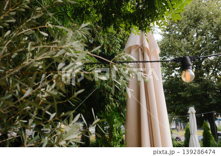 A closed beige umbrella stands next to hanging lights and green trees on a sunny day 139286474