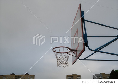 A basketball hoop and backboard are outside with buildings visible beneath a cloudy sky A basketball hoop and backboard are outside with buildings visible beneath a cloudy sky 139286564