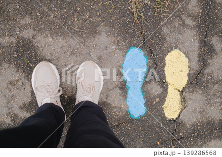 Person wearing shoes stands near blue and yellow painted footprints on asphalt Person wearing shoes stands near blue and yellow painted footprints on asphalt 139286568