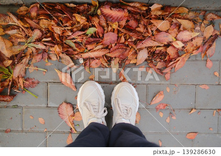 Feet in athletic shoes stand on a grey brick pathway covered in autumn leaves Feet in athletic shoes stand on a grey brick pathway covered in autumn leaves 139286630