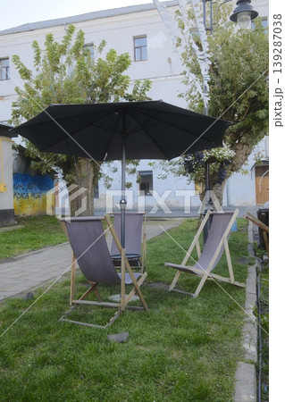 Several lounge chairs are set up under a large umbrella on grass near a building Several lounge chairs are set up under a large umbrella on grass near a building 139287038