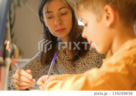 A mother helping her son with his studies. Homework assistance. Warm lighting. A cozy atmosphere for family learning. A focused student's pose. A scene of studying before bed. Space for text. Banner. 139289656