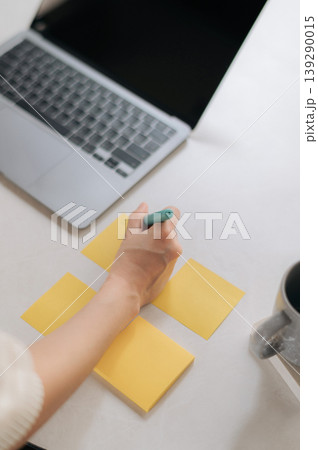 Dedicated young asian woman feeling highly productive writing business strategy for new mobile smartphone app on yellow sticky paper next to modern silver laptop upon bright white desk 139290015