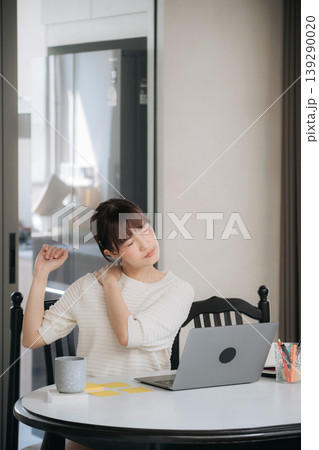 Exhausted young asian woman experiencing severe neck pain and shoulder stiffness after long business hours working on laptop computer and testing mobile app on smartphone sitting at round table 139290020