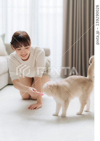 Joyful young asian woman takes relaxing break from business laptop work and smartphone app to happily feed fluffy pet cat on floor of bright comfortable living room Joyful young asian woman takes relaxing break from business laptop work and smartphone app to happily feed fluffy pet cat on floor of bright comfortable living room 139290026