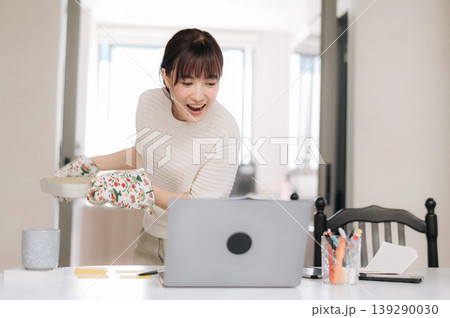 Excited young Asian woman holding hot baking dish with oven mitt looking surprised at laptop screen while managing home cooking business with smartphone app on kitchen table Excited young Asian woman holding hot baking dish with oven mitt looking surprised at laptop screen while managing home cooking business with smartphone app on kitchen table 139290030