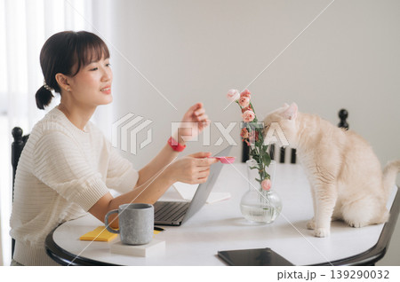 Cheerful young asian woman managing business on laptop and smartphone app smiling lovingly at cute pet cat smelling pretty flower on modern table in bright room Cheerful young asian woman managing business on laptop and smartphone app smiling lovingly at cute pet cat smelling pretty flower on modern table in bright room 139290032