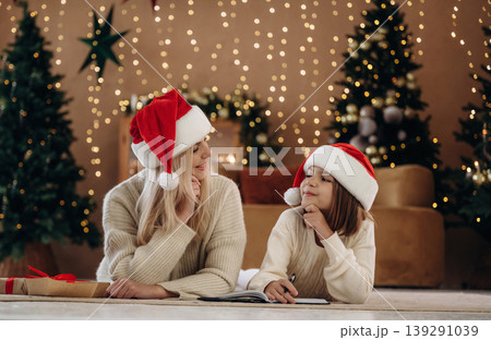 On the floor, lying down, holding pen, in Santa hats. Mother and daughter are celebrating New Year at home in domestic room On the floor, lying down, holding pen, in Santa hats. Mother and daughter are celebrating New Year at home in domestic room 139291039