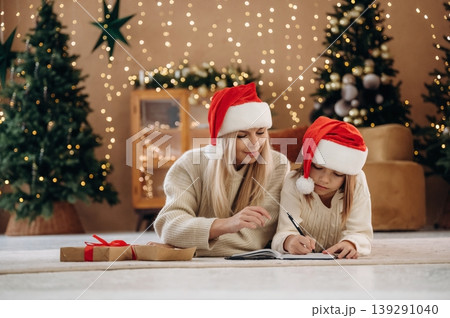 On the floor, lying down, holding pen, in Santa hats. Mother and daughter are celebrating New Year at home in domestic room 139291040