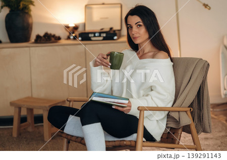 Beautiful brunette is with cup of tea. Relaxed woman is sitting in the chair and reading the book at home Beautiful brunette is with cup of tea. Relaxed woman is sitting in the chair and reading the book at home 139291143