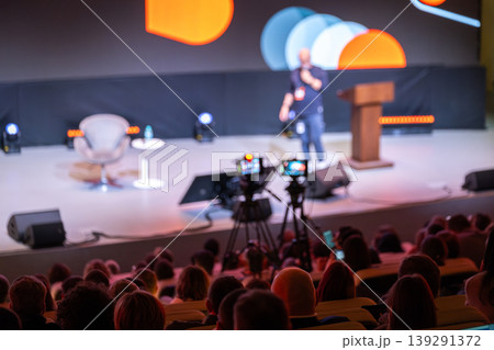Conference speaker on stage with audience and cameras filming live presentation at event 139291372