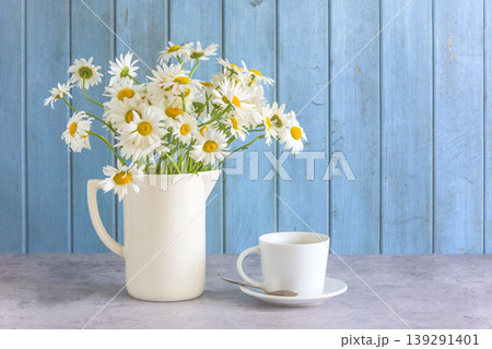Happy birthday or mother's day greeting card template; bouquet of white daisy flowers in white jug and white cup of tea or cofffee on a table nearr blue paint wooden background; copy space Happy birthday or mother's day greeting card template; bouquet of white daisy flowers in white jug and white cup of tea or cofffee on a table nearr blue paint wooden background; copy space 139291401