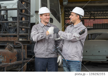 Caucasian and Middle Eastern mid-adult male mechanics fist bumping by truck in Asian industrial yard 139292355
