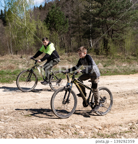 Father and son enjoy a bike ride on a sunny day, traversing a dirt path through a forest. Father and son enjoy a bike ride on a sunny day, traversing a dirt path through a forest. 139292359