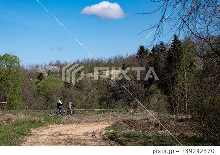 Two cyclists on a dirt road, surrounded by lush greenery and trees under a clear blue sky. Two cyclists on a dirt road, surrounded by lush greenery and trees under a clear blue sky. 139292370