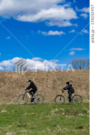 Two cyclists enjoy a sunny day riding mountain bikes across a grassy field under a vibrant blue sky. Two cyclists enjoy a sunny day riding mountain bikes across a grassy field under a vibrant blue sky. 139292457