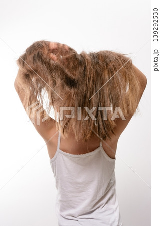 Girl with long brown hair, viewed from behind, holding her hair. She's wearing a white tank top against a white background. 139292530