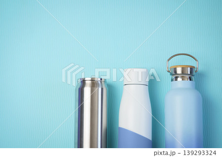 Three reusable drink bottles stainless can, insulated white bottle, and blue travel flask side by side on a textured aqua background, suggesting hydration, eco-friendly lifestyle, and modern design. Three reusable drink bottles stainless can, insulated white bottle, and blue travel flask side by side on a textured aqua background, suggesting hydration, eco-friendly lifestyle, and modern design. 139293324