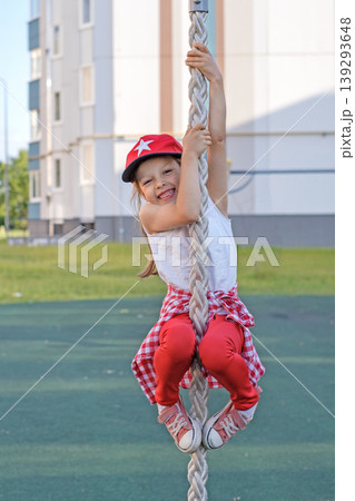 A funny laughing girl in a red cap climbs up a rope. Children's yard playground A funny laughing girl in a red cap climbs up a rope. Children's yard playground 139293648
