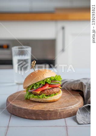 Meat burger with tomato, cheese, and lettuce on a wooden board on a table with glass of water Meat burger with tomato, cheese, and lettuce on a wooden board on a table with glass of water 139293821