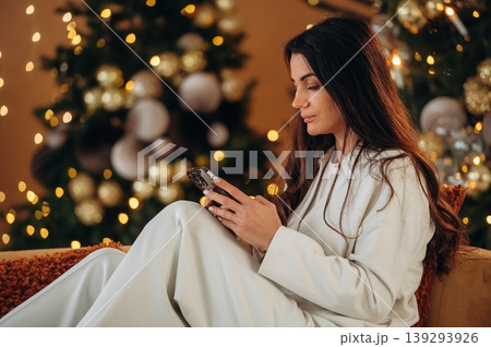 Smartphone is in hands, scrolling. Woman is sitting on the sofa in Christmas decorated room at New Year time celebration Smartphone is in hands, scrolling. Woman is sitting on the sofa in Christmas decorated room at New Year time celebration 139293926
