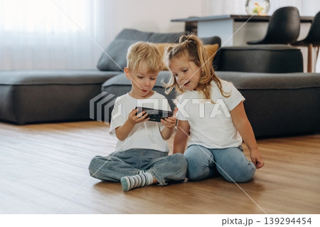 Smartphone in hands of boy. Little kids are playing together in the domestic room at daytime Smartphone in hands of boy. Little kids are playing together in the domestic room at daytime 139294454