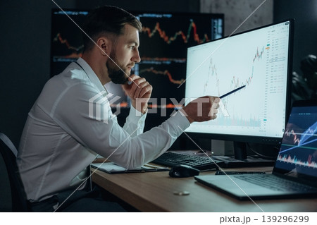View from the side, sitting and looking at displays. Handsome businessman is in the office with stock exchange market information on displays View from the side, sitting and looking at displays. Handsome businessman is in the office with stock exchange market information on displays 139296299