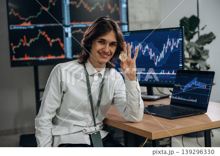Investment concept, holding coin of crypto currency. Young man is sitting in the office with multiple displays of stock market data 139296330