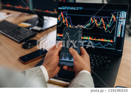 Against the laptop. Detailed close up view of man's hands that is holding smartphone with stock market crypto information Against the laptop. Detailed close up view of man's hands that is holding smartphone with stock market crypto information 139296830