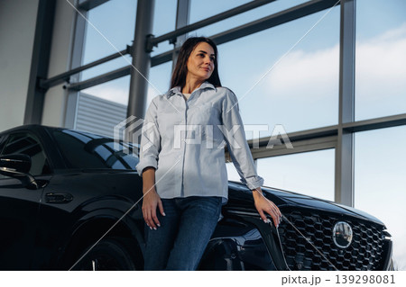 Big panoramic window is behind. Woman posing with a car in an auto showroom, modern dealership environment Big panoramic window is behind. Woman posing with a car in an auto showroom, modern dealership environment 139298081
