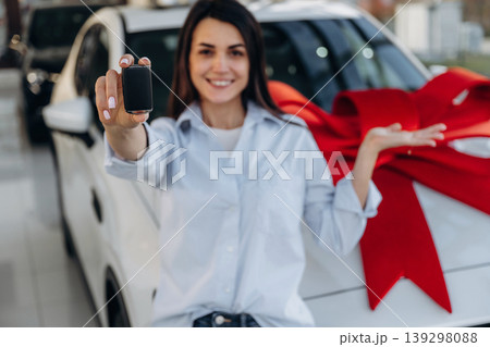 Red gift ribbon on a car. Woman posing with a car in an auto showroom, modern dealership environment 139298088