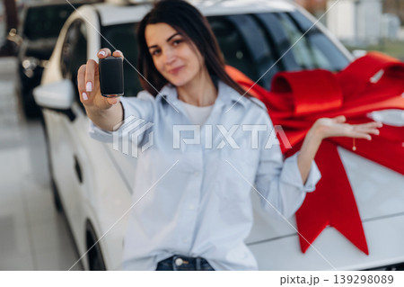 Red gift ribbon on a car. Woman posing with a car in an auto showroom, modern dealership environment Red gift ribbon on a car. Woman posing with a car in an auto showroom, modern dealership environment 139298089