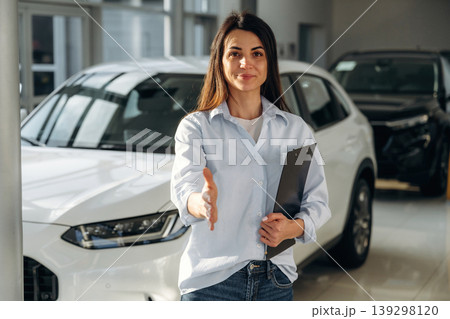 Giving hand to shake. Woman posing with a car in an auto showroom, modern dealership environment 139298120
