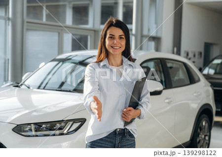 Giving hand to shake. Woman posing with a car in an auto showroom, modern dealership environment 139298122