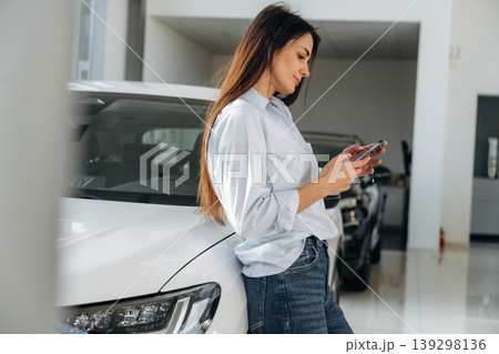 Smartphone is in hands, leaning on the vehicle. Woman posing with a car in an auto showroom, modern dealership environment Smartphone is in hands, leaning on the vehicle. Woman posing with a car in an auto showroom, modern dealership environment 139298136
