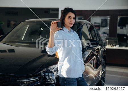In shirt and jeans, holding keys. Woman posing with a car in an auto showroom, modern dealership environment 139298437