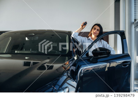 Out of the opened door, with keys in hand. Woman posing with a car in an auto showroom, modern dealership environment Out of the opened door, with keys in hand. Woman posing with a car in an auto showroom, modern dealership environment 139298479