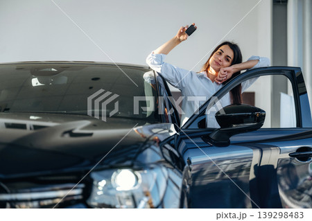 Out of the opened door, with keys in hand. Woman posing with a car in an auto showroom, modern dealership environment 139298483