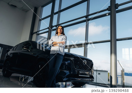 Big panoramic window is behind. Woman posing with a car in an auto showroom, modern dealership environment Big panoramic window is behind. Woman posing with a car in an auto showroom, modern dealership environment 139298488