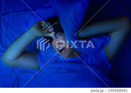 An angry man with a sleep mask covers his ears with a pillow, suffering from insomnia, noise, and sleep deprivation in a dark blue bedroom at night. An angry man with a sleep mask covers his ears with a pillow, suffering from insomnia, noise, and sleep deprivation in a dark blue bedroom at night. 139300091