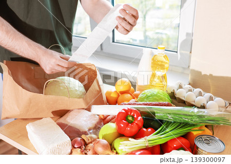 man checks a grocery receipt while unpacking fresh food from a paper bag, including vegetables, meat, and eggs on a kitchen table. man checks a grocery receipt while unpacking fresh food from a paper bag, including vegetables, meat, and eggs on a kitchen table. 139300097