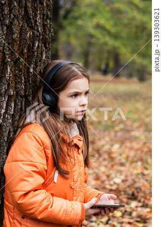 Young girl in orange jacket wearing headphones and holding smartphone by a tree in autumn park Young girl in orange jacket wearing headphones and holding smartphone by a tree in autumn park 139303621