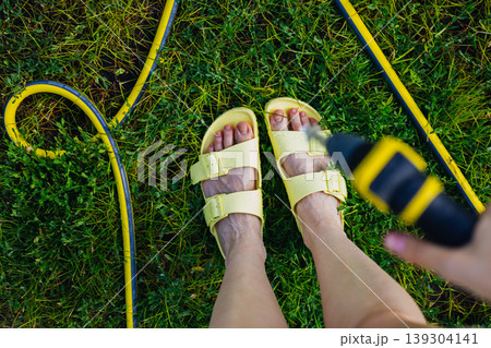 Woman in yellow sandals spraying water on her feet with a garden hose to cool down. Concept of summer heat relief, sensory wellness, and backyard fun. Authentic refreshing moment in a sunny garden. 139304141